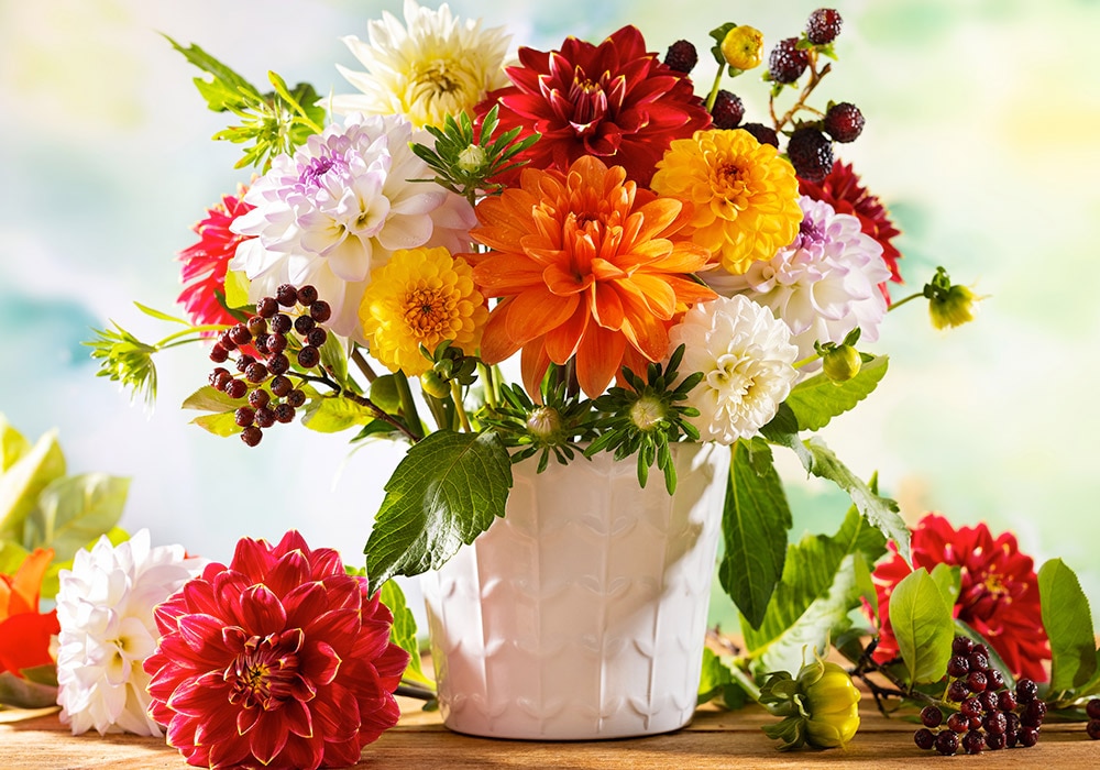 A white vase holding a colorful mix of flowers and berries, set on a wooden surface against a blurred, bright background. Various vibrant flowers surround the vase.